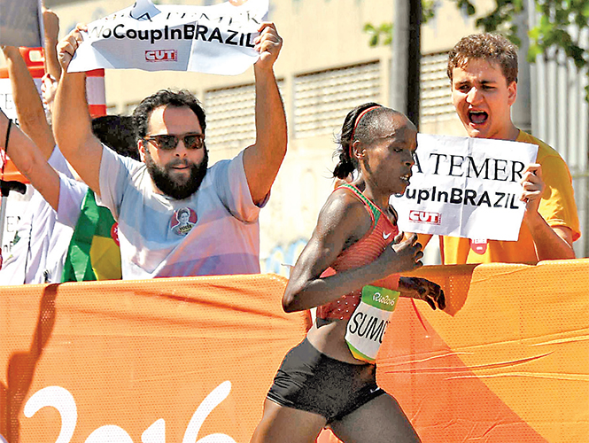 La corredora keniana Jemima Jelagat Sumgong corre en el maratón mientras un grupo de manifestantes muestra banderas que dicen: “Fora Temer” (Fuera Temer, presidente interino de Brasil). Foto: AFP